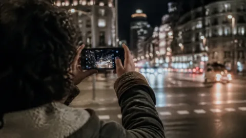 Pessoa fotografando uma rua iluminada à noite com celular
